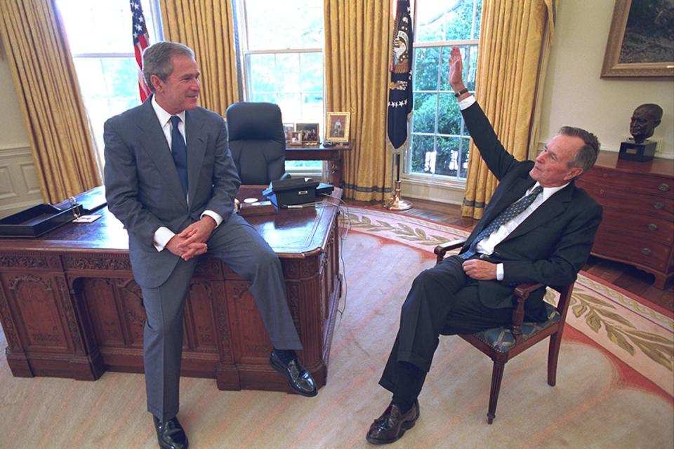 Two men, one sitting on a desk and the other in a chair, smile and gesture at each other