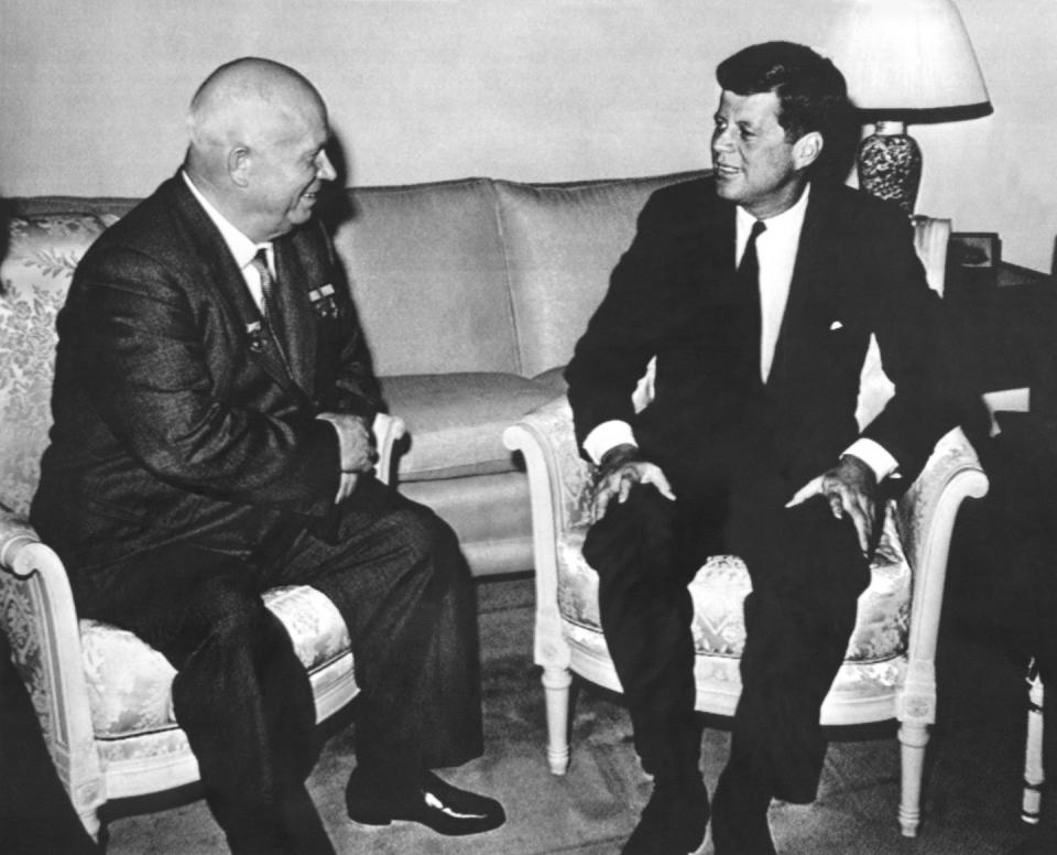 An elderly man with a balding head talks with a younger man as they sit near each in two chairs.