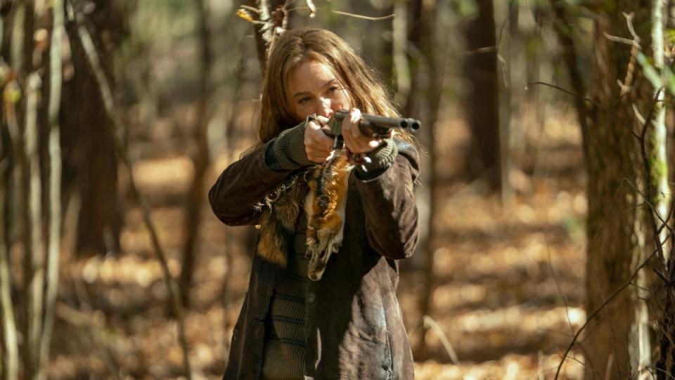 Actress Lynn Collins as Leah on The Walking Dead stands in a sparse forest with brown leaves on the ground holding up a shotgun at an unseen enemy