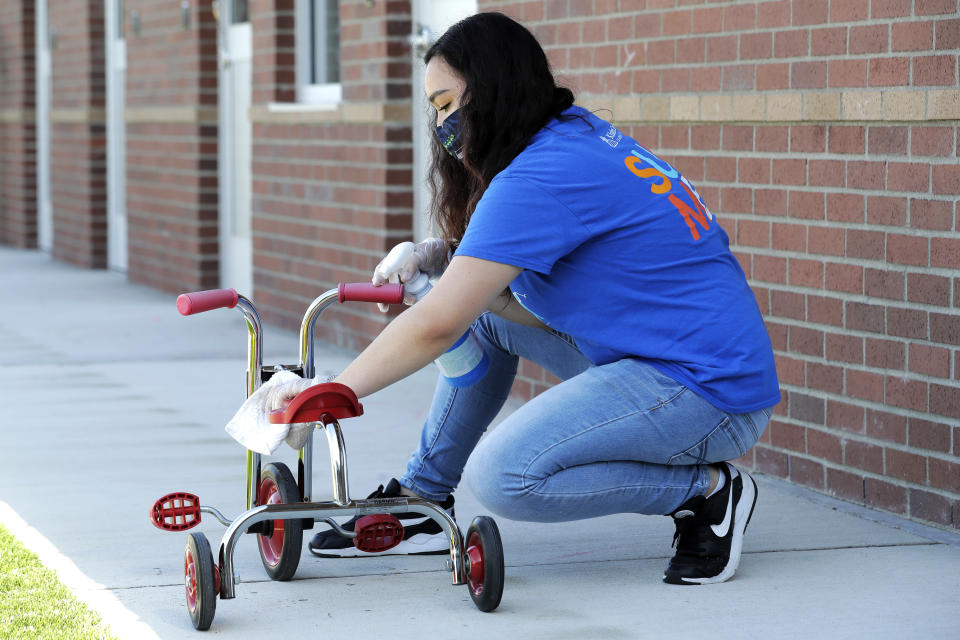 In this May 27, 2020 photo, Alena Kleinman, a worker at the Frederickson KinderCare daycare center in Tacoma, Wash., wears a mask as she cleans a tricycle following use by a class, a task that is repeated several times a day. In a world weary of the coronavirus, many working parents with young children are now struggling with the decision on when or how they'll be comfortable returning to their child care providers. Frederickson KinderCare has been open throughout the pandemic to care for children of essential workers. (AP Photo/Ted S. Warren)
