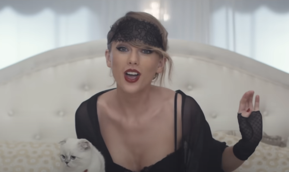 Taylor Swift in a black outfit with a mesh veil, posing on a bed with a white cat