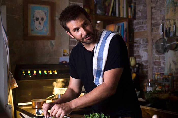 Man in a kitchen leaning over a counter with fresh herbs, cooking with a thoughtful expression