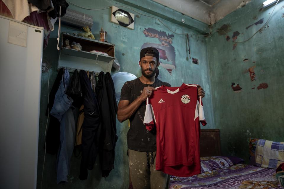 El futbolista Mahrous Mahmoud muestra la camiseta de la selección egipcia que usa en los entrenamientos el 9 de mayo del 2020 en su casa de Manfalut. Puntal del líder de la segunda división Beni Suef, Mahmoud está trabajando en un mercado mientras espera la reanudación del campeonato, suspendido por el coronavirus. (AP Photo/Nariman El-Mofty)