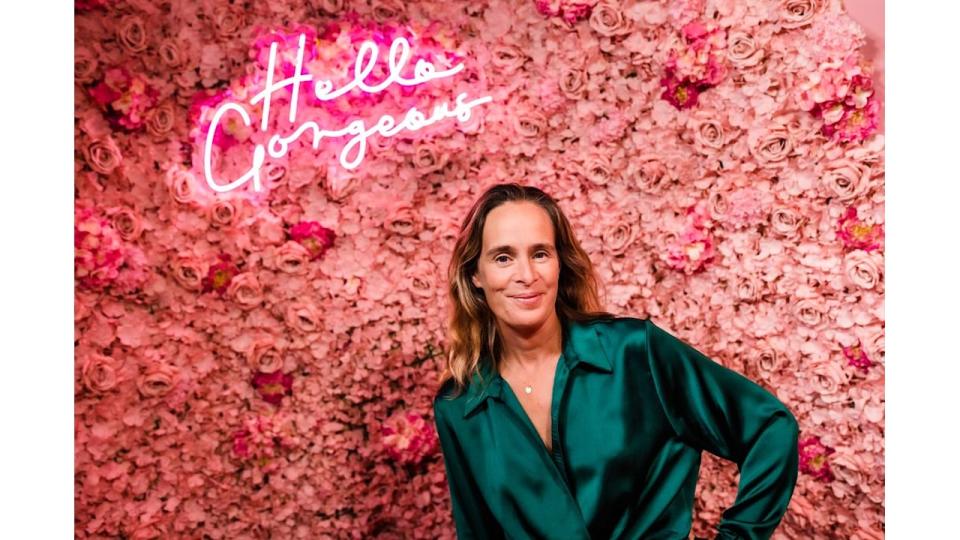 woman in a silky green shirt posing in front of a pink flower wall with hello gorgeous neon light