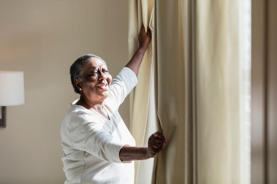 A senior woman at home at a window in her bedroom, opening the curtains to let the sunlight in.