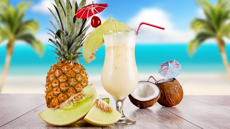 A garnished piña colada sitting next to a pineapple and coconut near a beach