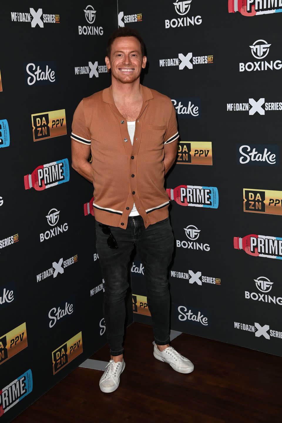 joe swash, a young man stands smiling at the camera and standing with hands behind his back, he wears black jeans with a brown tshirt