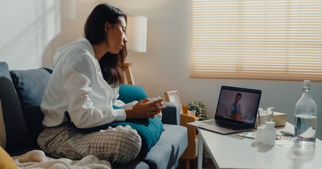 <p>MTStock Studio / Getty Images</p> Young Asian female using laptop to speak with her healthcare provider while sitting on sofa in living room at home.