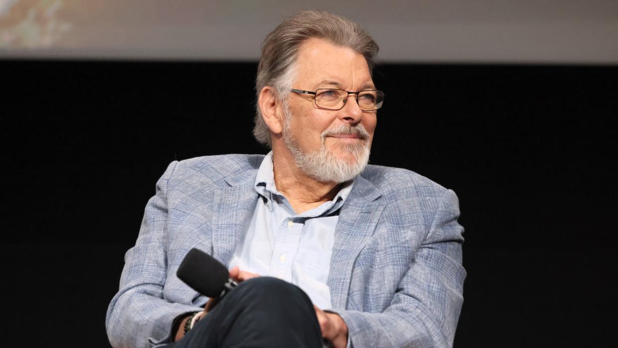  A bearded man in a grey suit sits on a stage holding a microphone. 