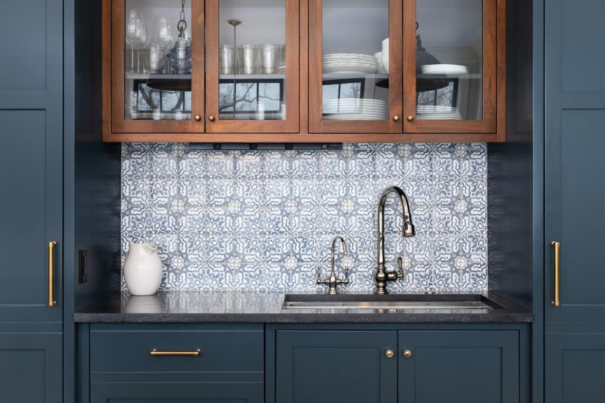 a kitchen sink with a beautiful pattern tiled backsplash with a chrome faucet, black granite countertops, and surrounded by blue and wood cabinets