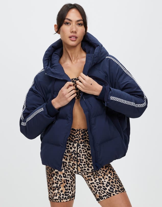 A woman stands in black and orange cheetah print active shorts and sports bra wrapped in an oversized navy puffer jacket with white sleeve highlights