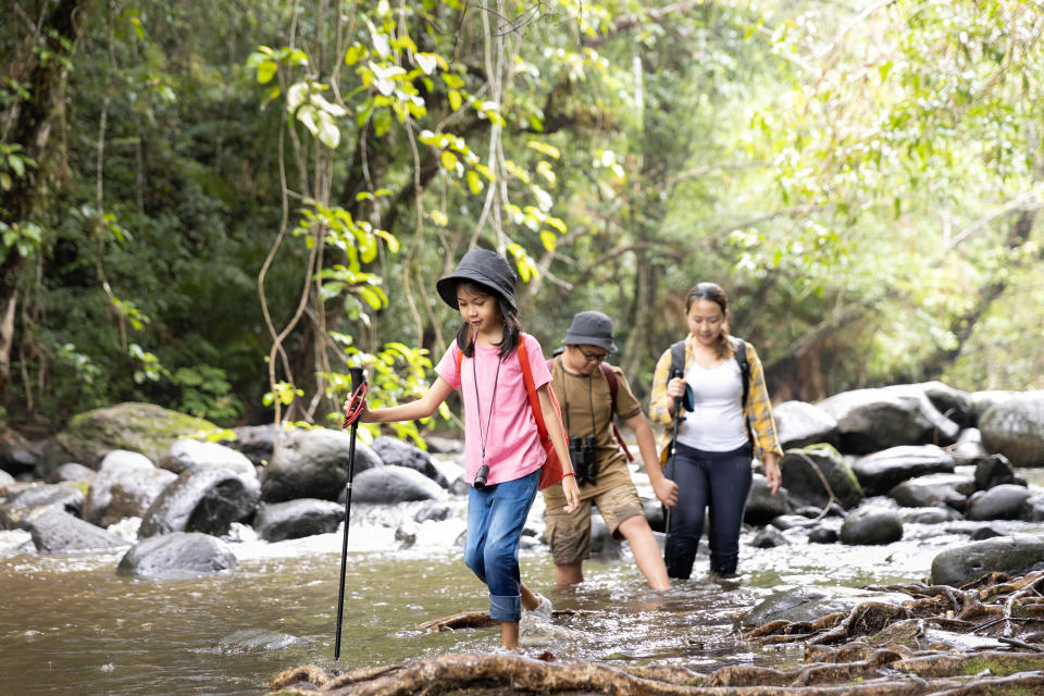 Family Asian, mother, son, and daughter, on a hiking adventure, walking crossing a stream flowing water in the forest, wearing a backpack and holding a hiking pole.