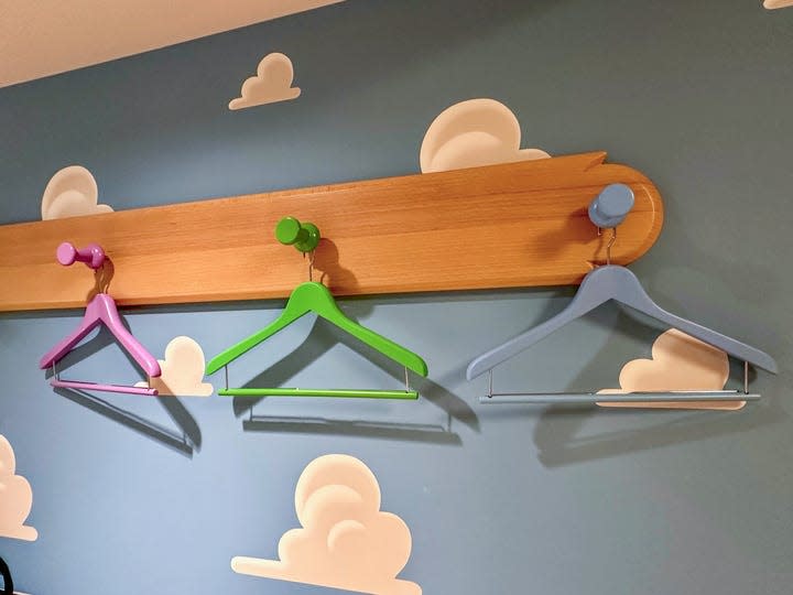 Three empty hangers, pink, green and light blue, hang on wooden pegs on a blue wall with white clouds in the Toy Story Hotel in Tokyo.