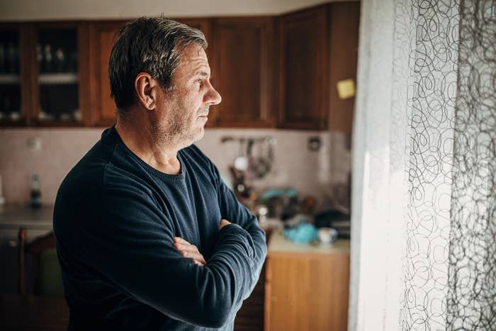 A man stands in a kitchen looking out the window with his arms crossed. The kitchen has wooden cabinets and various items on the counter