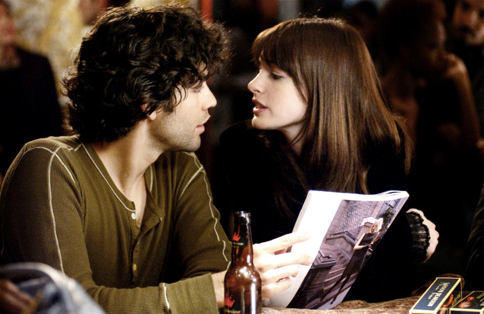 Two actors in a scene, man holding a book as woman gazes at him, intimate setting with a drink on the table