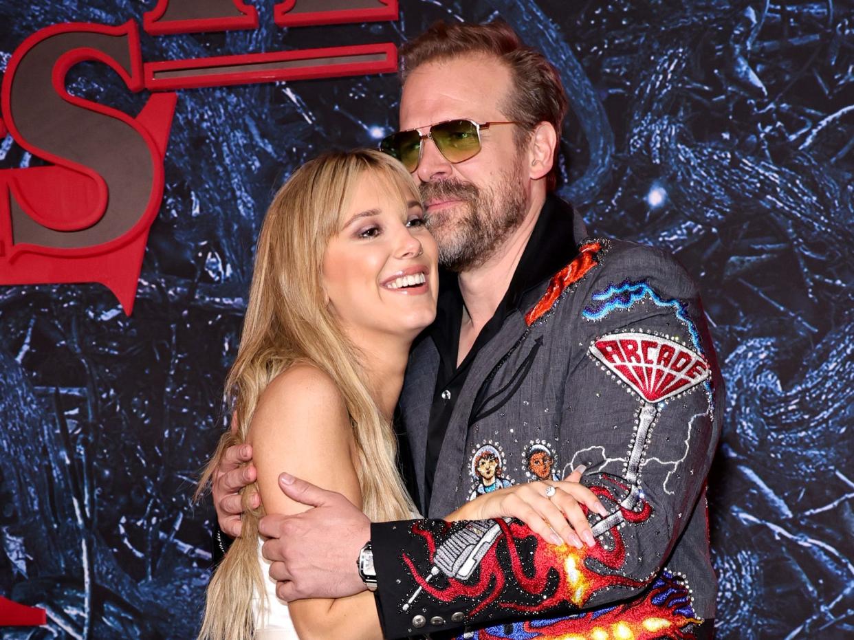 millie bobby brown with long blonde hair and a white gown, hugging david harbour wearing sunglasses and a black and grey jacket with space themed decals on the stranger things 4 red carpet