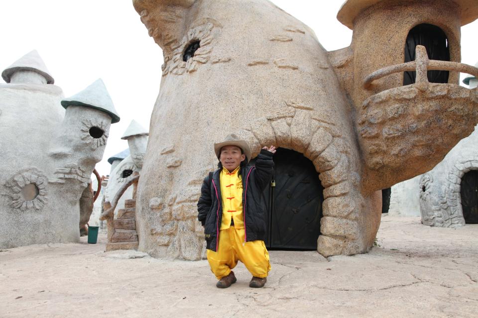 A person with dwarfism stands outside a fairytale style house at the