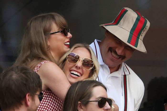 Taylor Swift, Brittany Mahomes, and Travis Kelce smile and hug each other outdoors. Taylor wears a gingham dress, Brittany wears sunglasses, and Travis wears a Gucci hat