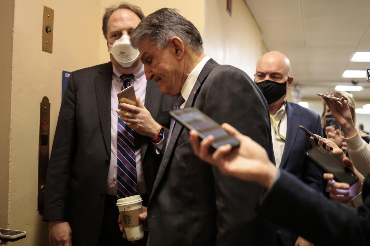 Sen. Joe Manchin, looking down and holding a cup of Starbucks coffee, is followed by reporters holding cellphones out toward him.