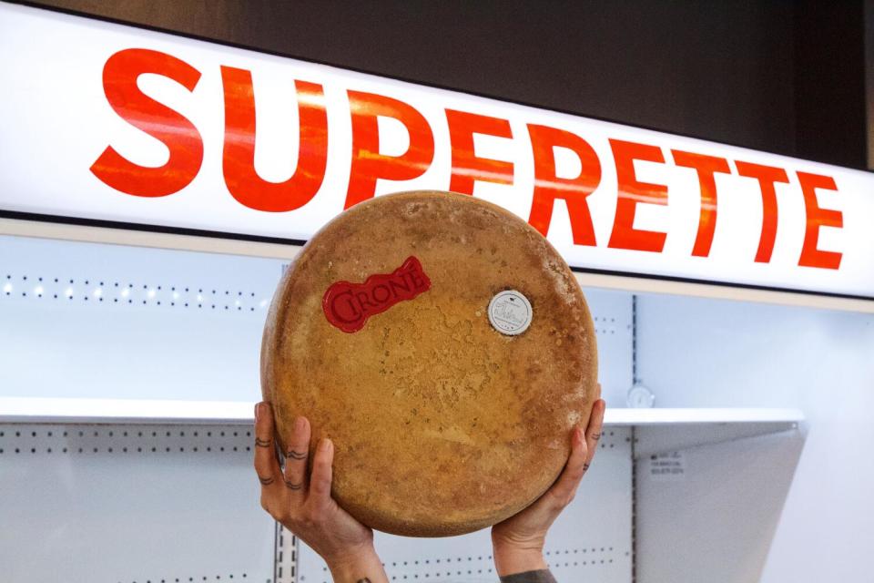 A hand holds up a wheel of cheese in front of a fridge that says "SUPERETTE" at DTLA Cheese Superette.