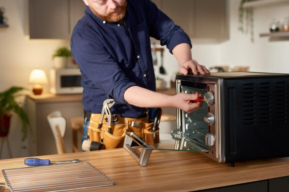 A man in a blue shirt uses a tool to fix a kitchen appliance. 