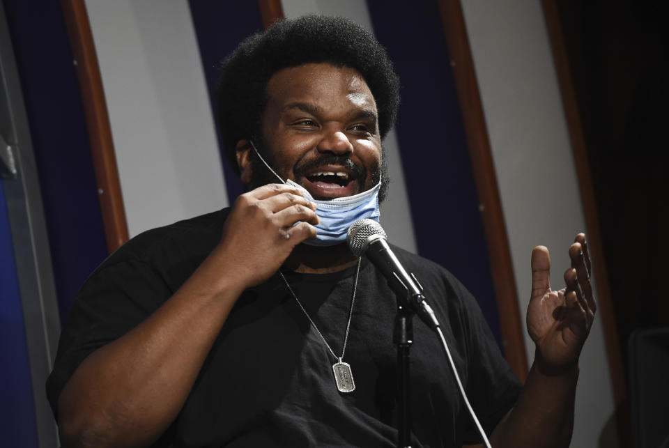 In this April 20, 2020, photo, comedian Craig Robinson lowers his mask as he performs during a "Laughter is Healing" stand-up comedy livestream event at the Laugh Factory comedy club in Los Angeles. With comedy clubs closed and concert tours put on hold, comics like Robinson, Tiffany Haddish, Will C and others are keeping the jokes flowing on webcasts and Zoom calls even without the promise of a payday, because they say the laughs are needed now more than ever. (AP Photo/Chris Pizzello)