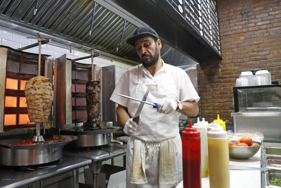 In this Wednesday, Oct. 9, 2019, photo, chef Diaa Alhanoun sharpens his knives between serving customers, while skewered pieces of cooked chicken and beef known, or "shawarma," are kept warm at his restaurant Sakib, in New York. A Syrian refugee fleeing civil war at home, Alhanoun, his wife and four children arrived in the U.S. from Jordan in 2016. Less than three years later Alhanoun and a partner opened a small restaurant in Brooklyn's trendy Williamsburg neighborhood. (AP Photo/Kathy Willens)