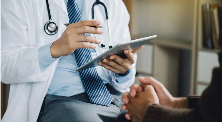 A doctor sits with a patient and takes notes on a tablet.