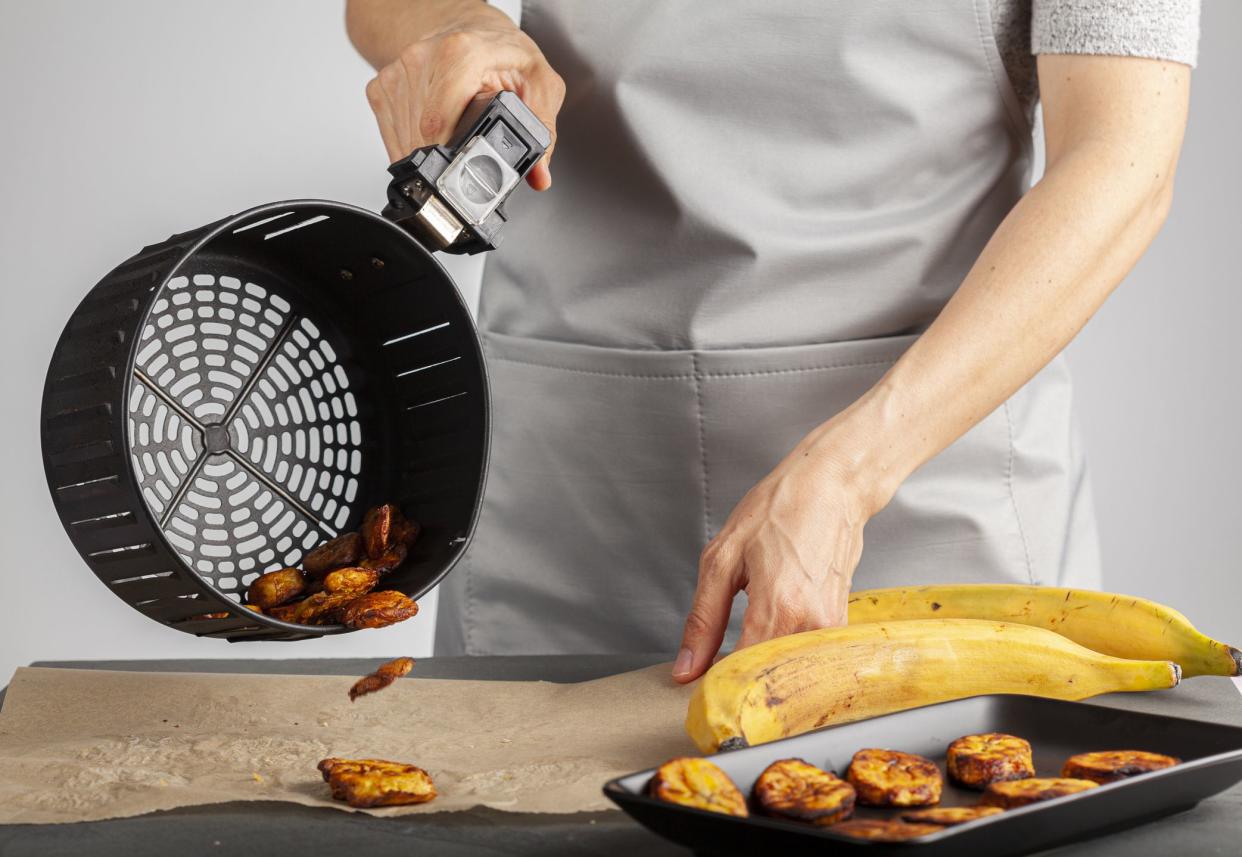 A caucasian woman is pouring freshly made plantain chips (Platanos Maduros) onto baking paper for cooling. She holds the handle of air fryer basket. Air fryer makes it low calorie compared to regular.