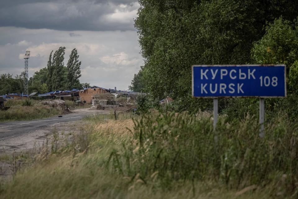 A blue road sign announcing the distance to the Kursk region of Russia among foliage and trees next to a gray road with a gray sky and a damaged structure in the background