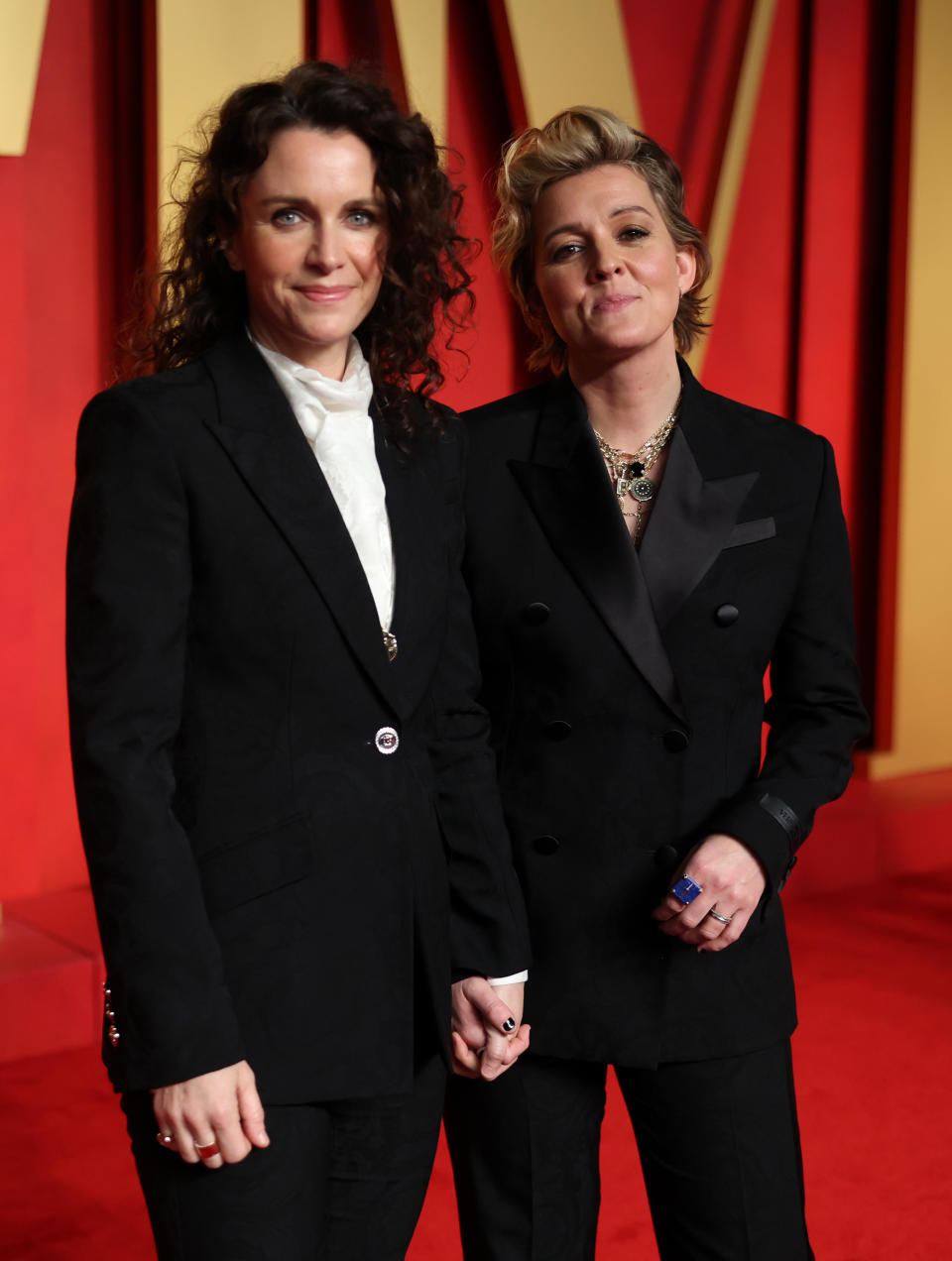 Brandi Carlile and Catherine Shepherd at a red carpet event, both wearing black suits with white shirts, posing together