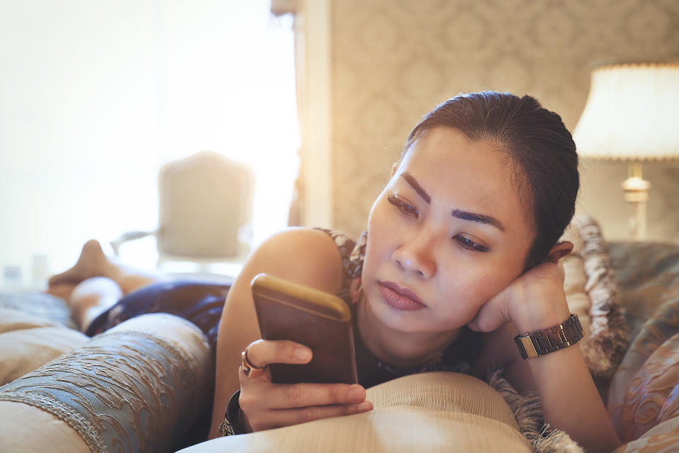 A woman lies on a bed propped on her elbows, looking intently at a smartphone screen, with her hand supporting her head