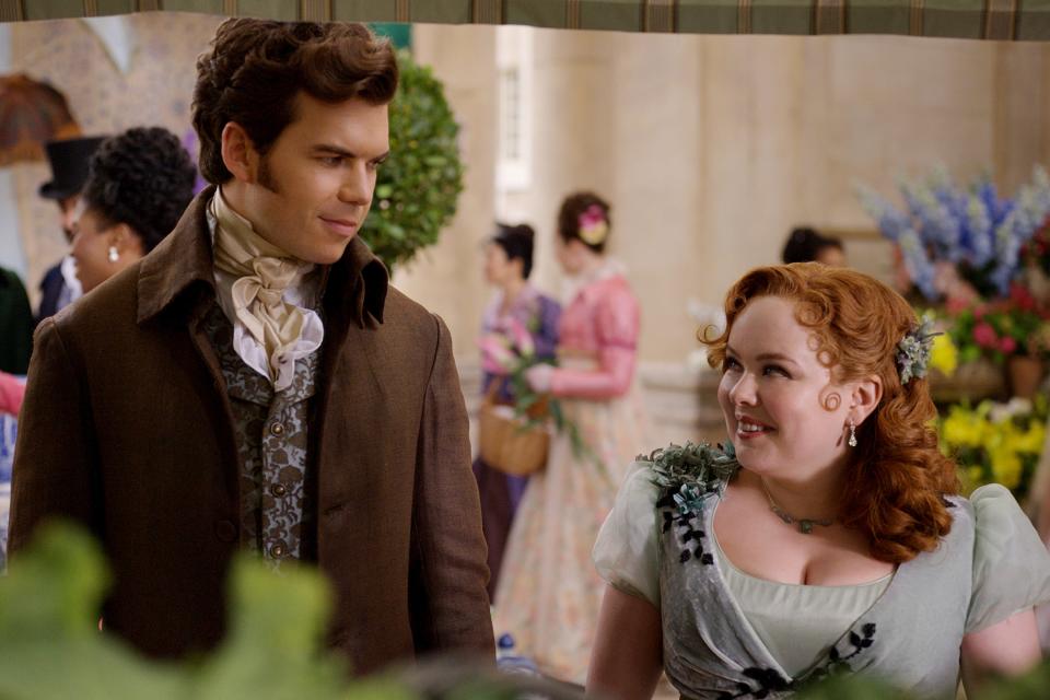 colin bridgerton and penelope featherington, played by luke newton and nicola coughlan, standing together and smiling at each other in a floral market