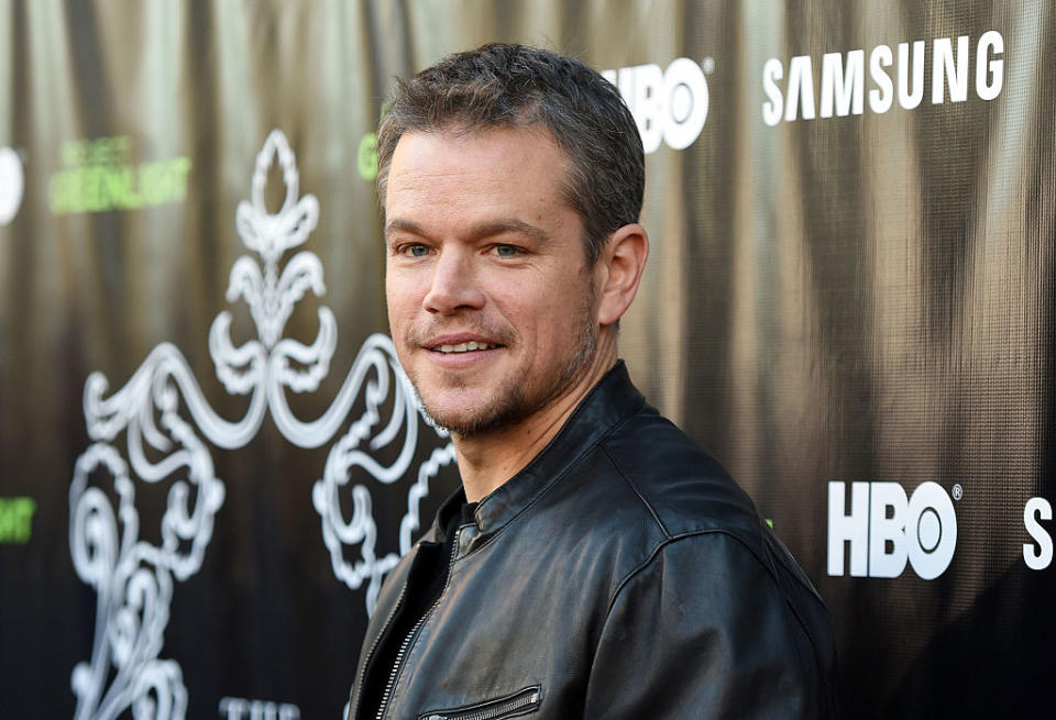 Matt Damon smiles in a leather jacket on the red carpet, with logos of HBO and Samsung in the background
