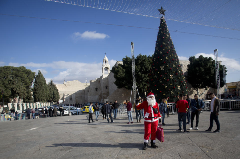 In this Thursday, Dec. 5, 2019, photo, a Palestinian wearing a Santa Claus costumes welcomes Christian visitors outside the Church of the Nativity, traditionally believed by Christians to be the birthplace of Jesus Christ, in the West Bank city of Bethlehem. As visitors descend on Bethlehem this Christmas, they have the option of staying in restored centuries-old guesthouses, taking food tours of local markets, and perusing the dystopian art in and around a hotel designed by the British graffiti artist Banksy.(AP Photo/Majdi Mohammed)