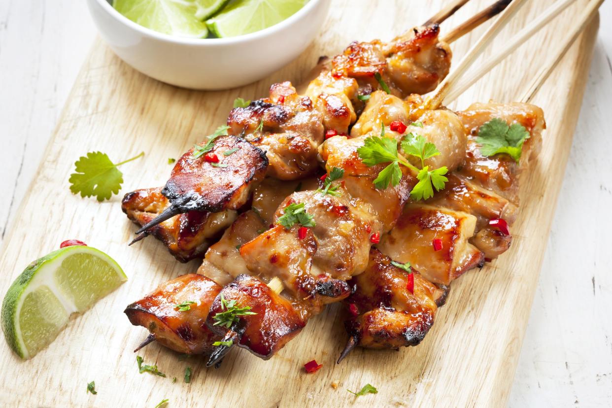 Five chicken satay with peanut sauce skewers with limes on a wooden cutting board on a white table