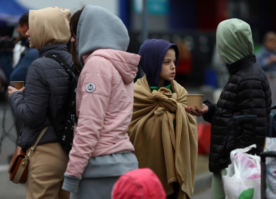 A young child wearing a knotted shawl looks backward as a crowd of refugees line up.