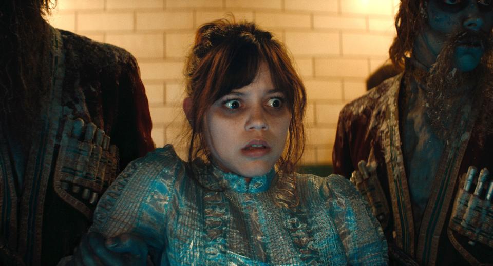 Jenna Ortega, in a period costume with intricate details, looks terrified as she stands between two undead figures in elaborate outfits in a scene from her television show