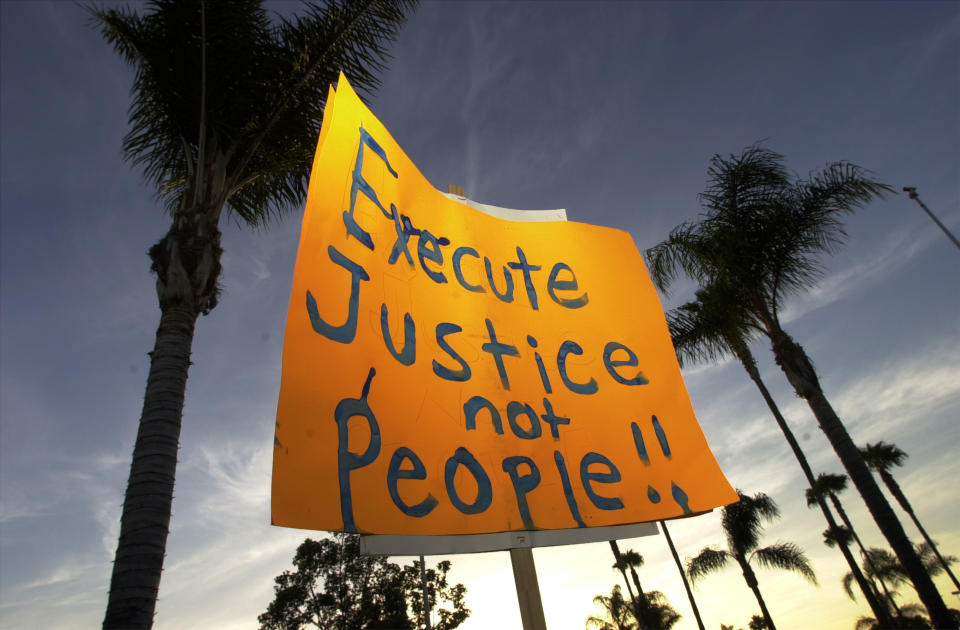 A hand-painted sign that reads Execute justice not people!! is seen in front of palm trees lit from behind by what appears to be a darkening sky.