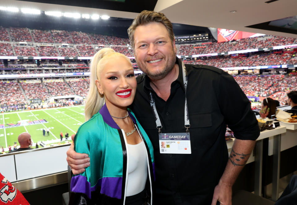 Gwen Stefani wears a teal and purple jacket next to Blake Shelton who's wearing a black Polo shirt