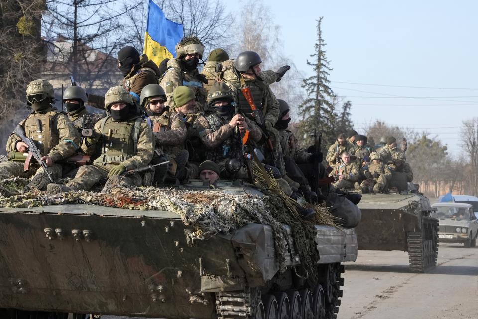 Two tanks loaded with soldiers drive along a road. A Ukrainian flag is displayed on the first tank.