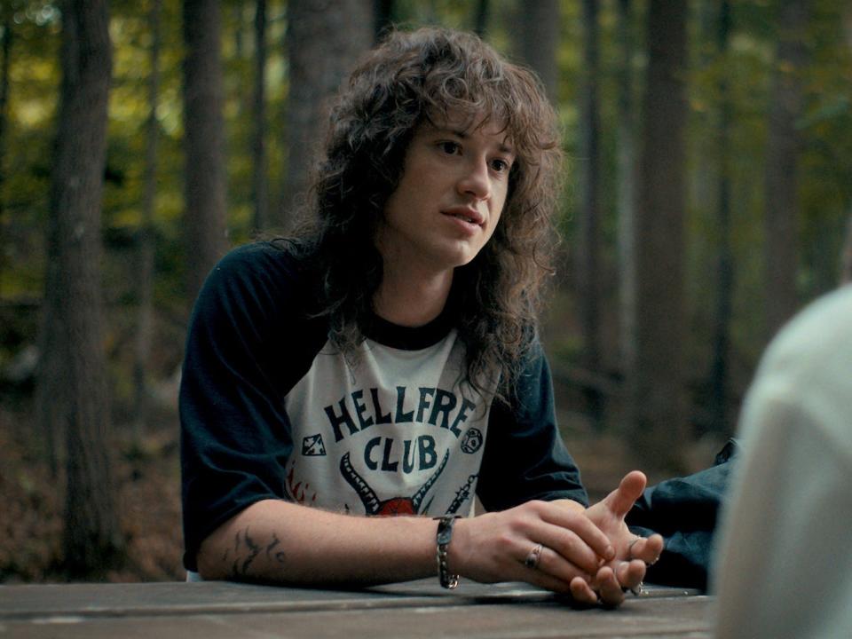 A young man with long curly hair and tattoos sits at a picnic table.