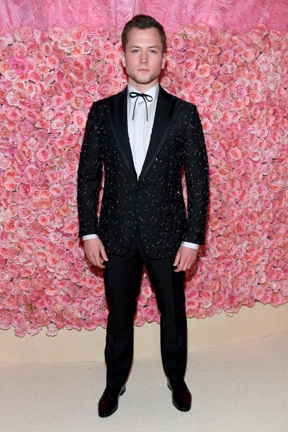 Taron Egerton stands in a black, sparkly tuxedo in front of a wall of pink flowers.