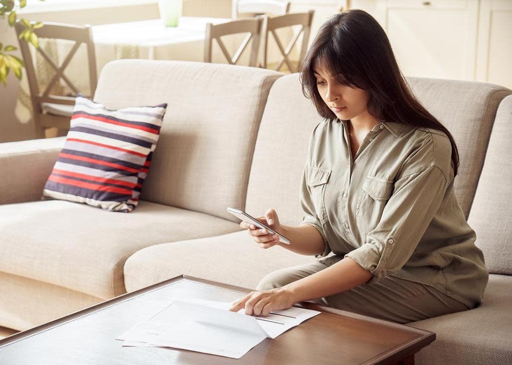 Woman holds smartphone in one hand while checking paperwork on table with the other.