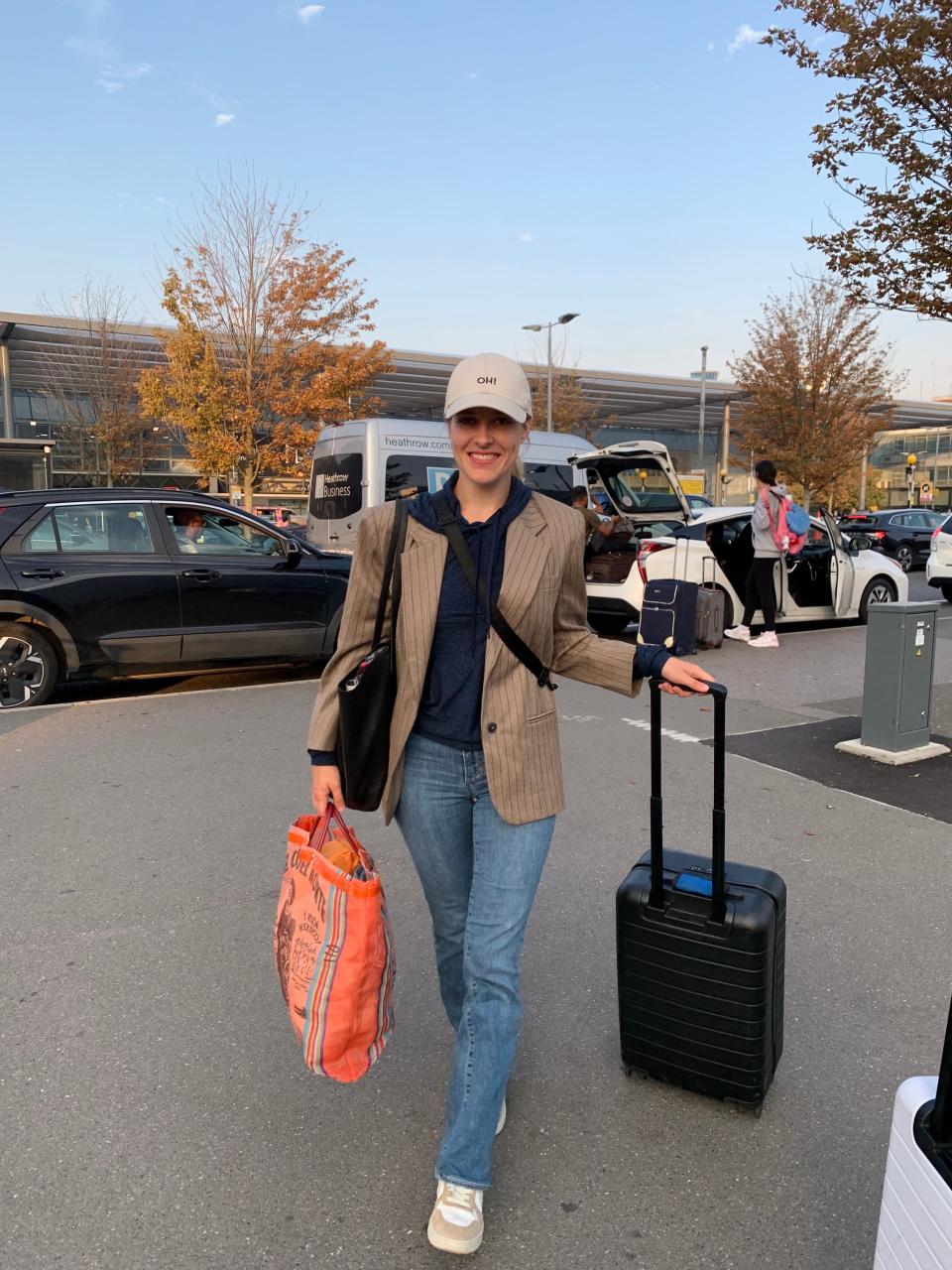 A woman standing in front of the airport with rolling luggage.