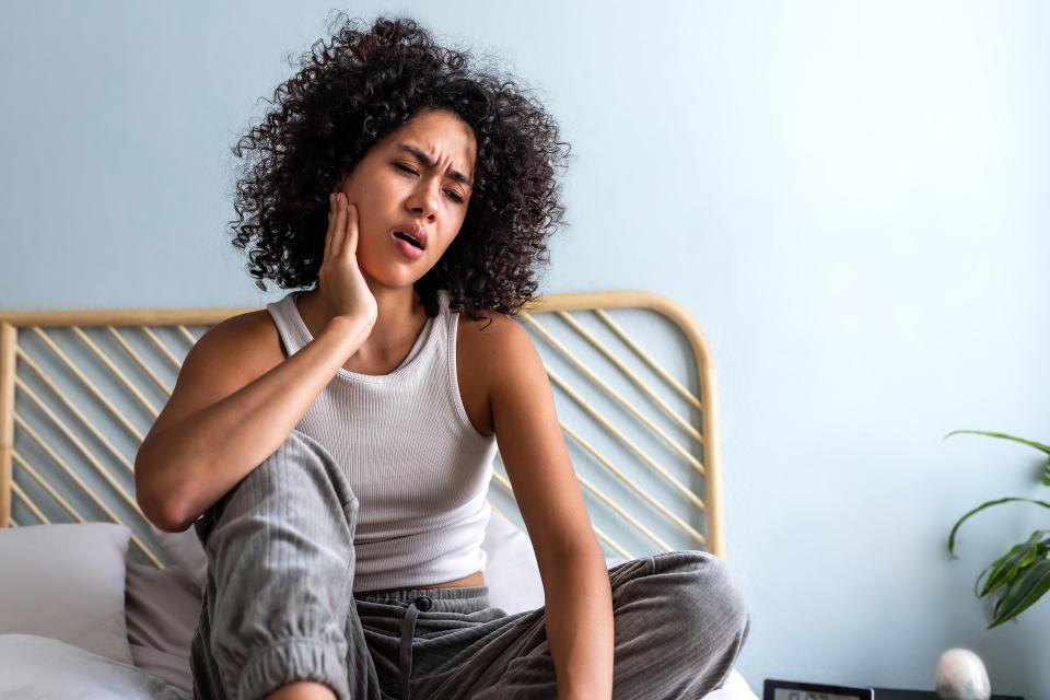 Young latina female with curly hair touching cheek in pain having toothache sitting on bed in cozy bedroom at home. Body concept.