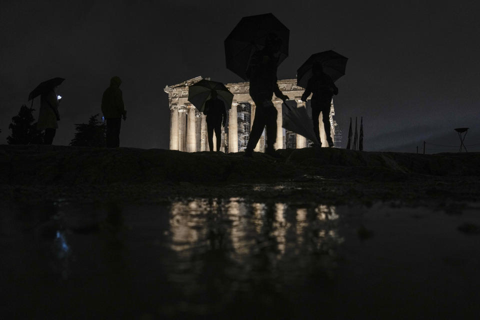 People with umbrellas walks past the ancient Parthenon temple at the Acropolis hill as they await the arrival of the Olympic flame as rain falls in Athens, Friday, April 19, 2024. The flame that will burn at the summer Olympics is spending the night at the ancient Acropolis in Athens, a week before its handover to Paris 2024 organizers. (AP Photo/Petros Giannakouris)