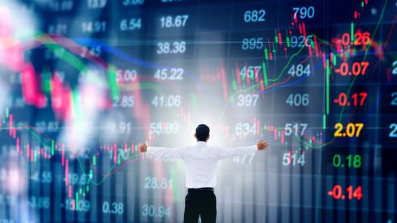 Man with arms outstretched in front of large display showing stock data and stock chart going up