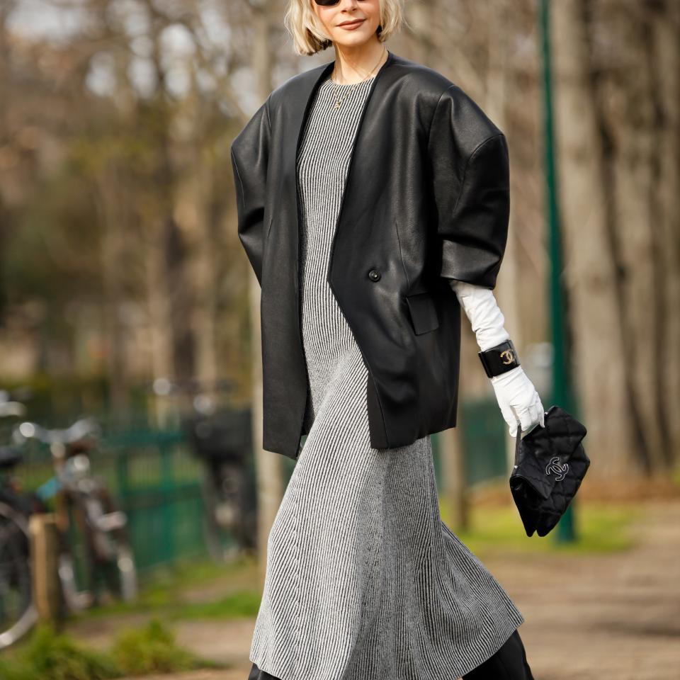 Model walking outdoors in a fitted ankle-length dress with an oversized blazer, black boots, sunglasses, and white gloves, holding a small clutch