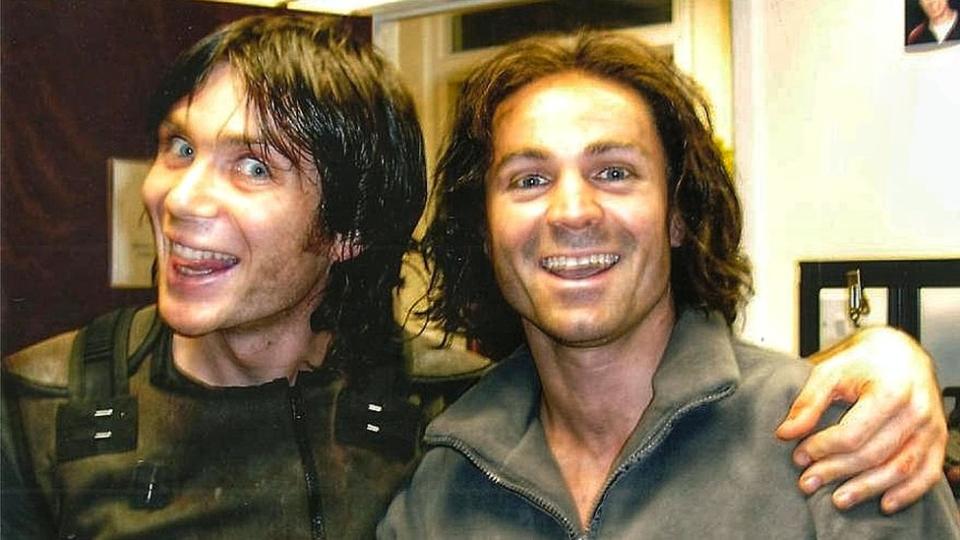 Nicholas Daines alongside actor Cillian Murphy on the set of Sunshine in 2007. Cillian (L) is a white man with straight dark hair just past his ears and bright blue eyes. He tilts his head to the right and smiles, mouth wide. Nicholas is styled to look like Cillian - they are the same height with the same hair and both wear green overalls and have make-up to make them look dirty. They're pictured inside in a cream-coloured room.